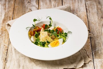 Close-up gourmet fish salad. Cod fillet, vegetables, avocado pate and fresh herbs. Restaurant serving. Chef's dish. Healthy and tasty food on a light wooden background. Fish dishes.