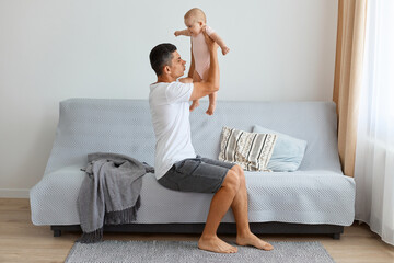 Indoor shot of ghandsome brunette father holding infant baby girl in hands and understand they need changing baby's diaper, feeling bad smell, raising kid up air.