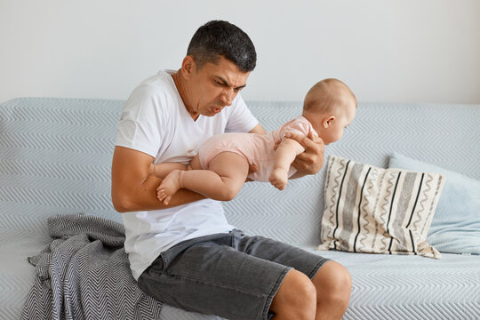 Portrait Of Frustrated Man Posing With Toddler Girl In Hands, Smelling Baby, Needs To Change Diaper And Wash Kid, Sitting On Sofa In Light Living Room At Home.