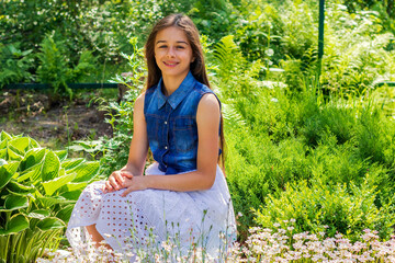 A cute happy girl in a summer dress with dark long hair sits in a summer sunny garden. 