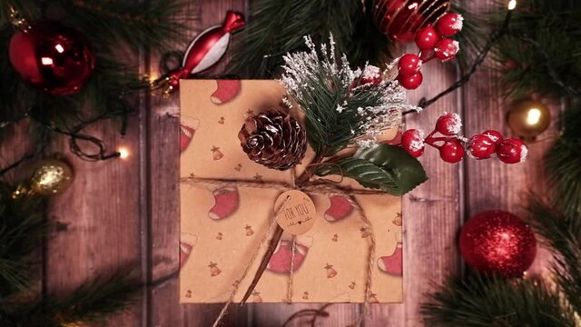 Putting a label "FOR YOU" on a Christmas gift box. Wooden background with fir branches, red balloons, toys for the Christmas tree. The garland glows with a warm light. POV. Top View 4K