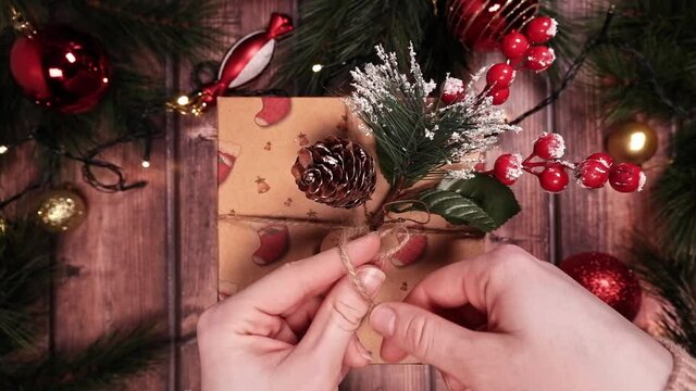 Tying a knot of twine on a Christmas gift box. Wooden background with fir branches, red balloons, toys for the Christmas tree. The garland glows with a warm light. POV. Top View 4K