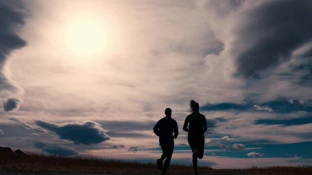 Striking black silhouette outline of a male and female jogger, jogging against a dramatic sky.  The joggers run away from the camera from left to right. The sun flares around the outline.