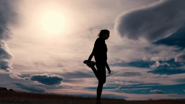 Beautiful black silhouette outline of a female jogger stretching her legs before a run.  She alternates legs stretching each quad or thigh individually.  A dramatic sky frames her.