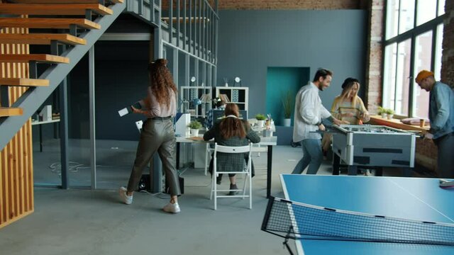 Young ambitious entrepreneurs are working in shared office while colleagues are playing foosball having fun. Coworking place and business activities concept.