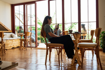 Young woman in casual shirt sitting at table in cafe and checking messenger while using smartphone.Full body shot in cafe interior.