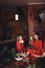 Two young women in red pajamas drinking coffee in cottage, december morning, christmass decoration