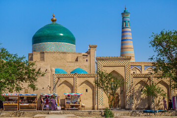 Fototapeta premium Two of main attractions of Khiva (Uzbekistan): Islam Khodja minaret and Pahlavan Mahmoud mausoleum with unique green dome. Merchants sell and tourists walk by walls below