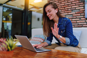 Cute young happy modern casual female wearing white headphones using laptop for video call,...