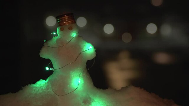 Little snowman litted by LED with pea eyes and bottle metal cap on head standing on snowdrift when night and snowing. Snowman is on the left of the frame, background is dark, out of focus with bokeh