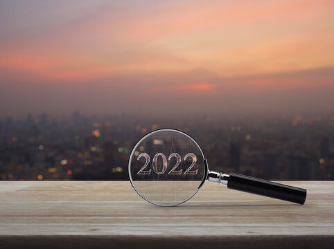 Magnifying glass with 2022 text and financial graph on wooden table over blur of cityscape on warm light sundown, Business happy new year 2022 research concept