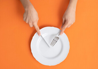 Female hands hold cutlery over the empty plate on orange background.