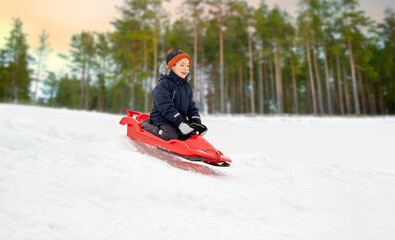Fototapeta na wymiar childhood, sledging and season concept - happy little boy sliding on sled down snow hill outdoors in winter over snowy forest or park background