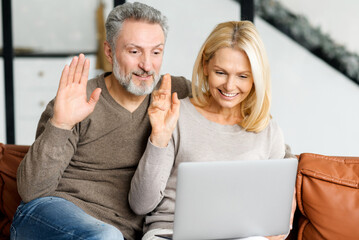 Lovely mature spouses using laptop computer for online communication, video connection with friends or family on the distance. Happy middle-aged couple sits on sofa, looks at the webcam and waving