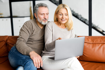 Cheerful middle-aged couple with a laptop sitting on the comfortable leather couch at home, choosing new furniture, making food order, scrolling news feed. Mature spouses spend leisure time together