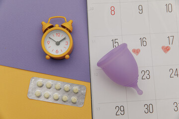 Calendar, alarm clock and menstrual cup with pain relief pills close-up. Woman critical days concept