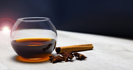 A cognac glass with a drink, a cinnamon stick, anise stars, on a wooden table, black background,...