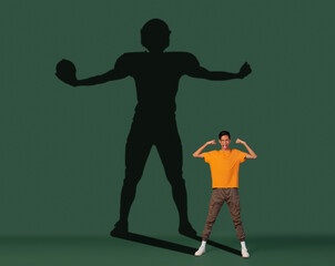 Fototapeta na wymiar Conceptual image with young boy dreaming about future sport career and shadow of fit male football player on dark green background