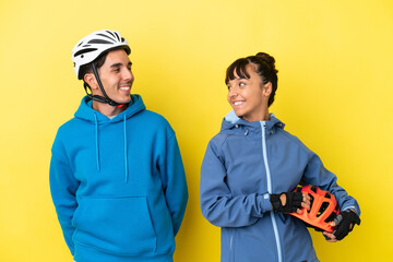 Young cyclist couple isolated on yellow background looking over the shoulder with a smile