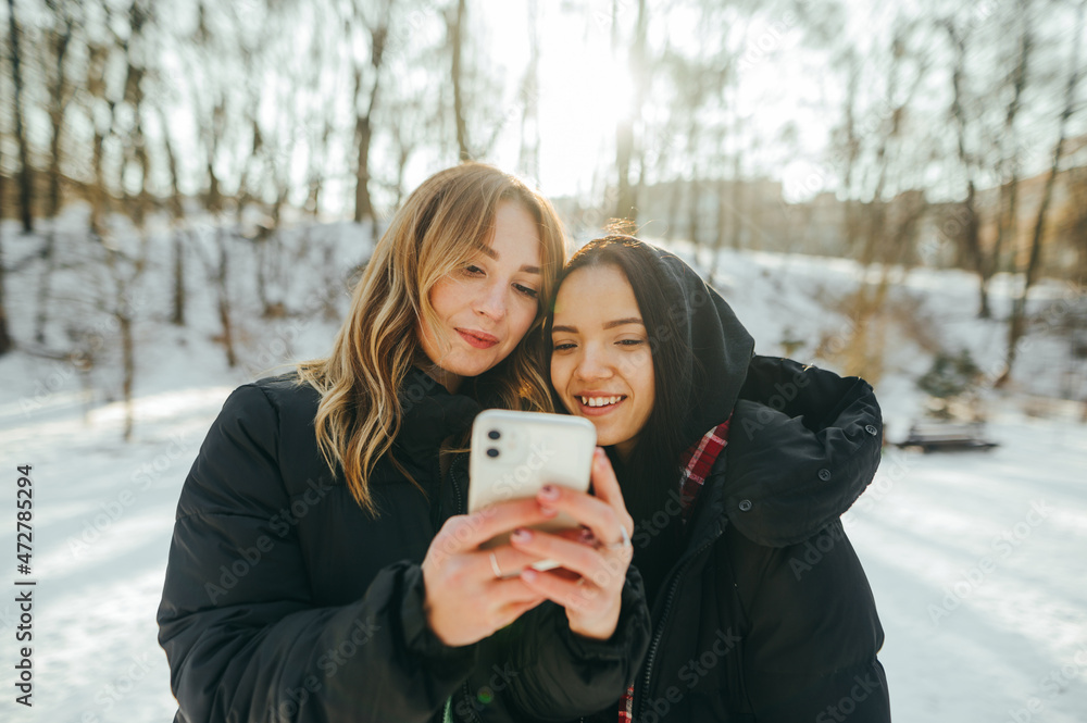 Wall mural Two beautiful women with smiles on their faces watching photos on a smartphone while walking in a snowy forest in the winter season.