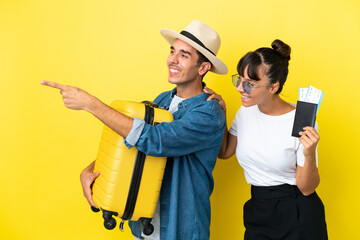 Young traveler friends holding a suitcase and passport isolated on yellow background pointing to...