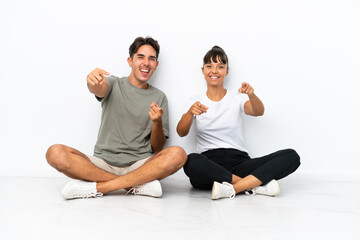 Fototapeta na wymiar Young mixed race couple sitting on the floor isolated on white background points finger at you while smiling