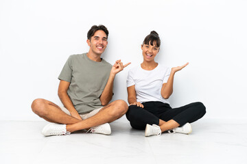 Fototapeta na wymiar Young mixed race couple sitting on the floor isolated on white background pointing finger to the side and presenting a product