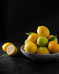 Citruses (lemons, limes and tangerines) in a gray plate on a black background. Nearby lies a cut half of a lemon with a leaf