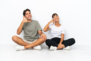 Fototapeta na wymiar Young mixed race couple sitting on the floor isolated on white background making phone gesture. Call me back sign