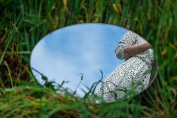 An oval mirror lies on the grass and reflects the waist of a woman in a beautiful dress. A...