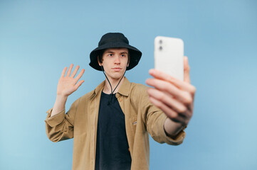Young man in a panama makes a video call to a smartphone on a blue background, looks at the...