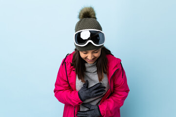 Mixed race skier girl with snowboarding glasses over isolated blue background smiling a lot.