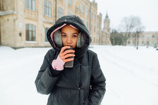 Positive woman in warm clothes and a hood drinking coffee from a cup under a chach walking around the town in the snow, looking at the camera with a happy face