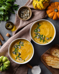 Two bowls of pumpkin soup with olive oil, cream and pumpkin seeds with bruschetta on gray background with linen napkin