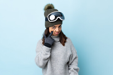 Mixed race skier girl with snowboarding glasses over isolated blue background showing something.