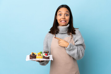 Pastry chef holding a big cake over isolated blue background frightened and pointing to the side.