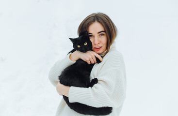 Positive lady in a knitted sweater poses for the camera on a background of snow with a black cat, looks at the camera and smiles. Winter. walk with a cat.