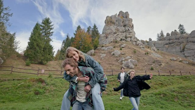 Five Young People Having Fun In Nature. Man With Beard Carries Girl On His Back. Others Run And Laugh. One Girl Taking Selfie Using Phone.