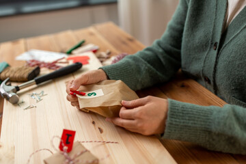 winter holidays and hobby concept - hands with clothespin, craft paper bag and tag making advent calendar on christmas at home