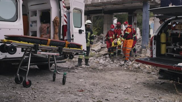 Multiracial team of paramedics in uniform and masks carrying spinal board with man and putting survivor on wheeled carrier near emergency vehicle during rescue mission on remains of destructed