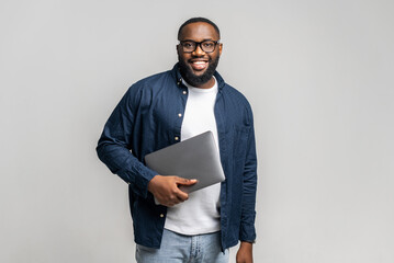 Smiling young African-American male entrepreneur stands in studio isolated on gray and carrying trendy laptop. Cheerful black guy in glasses and casual shirt looks at camera, holding closed laptop - Powered by Adobe