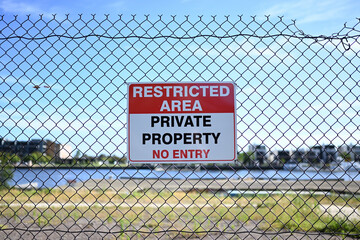 A restricted area sign, signalling that beyond the fence is private property and that no entry is permitted, on a fence. The undeveloped waterfront land can be seen in the background