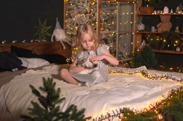 little beautiful blonde girl is sitting on a bed with a rabbit. Christmas mood. There is a Christmas tree and decorations in the background. The concept of holidays. Love for pets