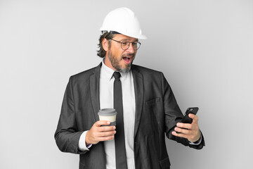 Young architect man with helmet over isolated background holding coffee to take away and a mobile