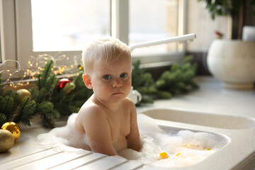 A blonde-haired baby is bathing in the sink in the kitchen against the background of a Christmas...