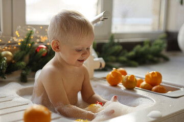 A blonde-haired baby is bathing in the sink in the kitchen against the background of a Christmas tree with garlands and oranges