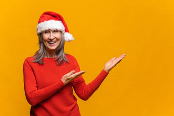 Fototapeta na wymiar Cheerful and happy senior woman in santa hat points with palms of hands to the empty space, excited mature lady happy to presenting novelty, copy space, mockup, isolated on the yellow
