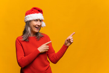 Fototapeta na wymiar Positive elderly woman in red santa hat looks and points to the side with both index fingers, isolated over yellow background, copy space, advertises or recommends a new product, holiday sale