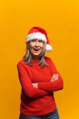 Joyful senior gray-haired woman in santa hat stands with arms crossed and looks at the camera, positive mature middle-aged lady smiles, celebrating new year and christmas isolated on yellow background