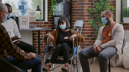 Woman with disability attending aa group therapy session with people in circle. Adult sitting in wheelchair and talking about alcohol addiction to patients during coronavirus pandemic.